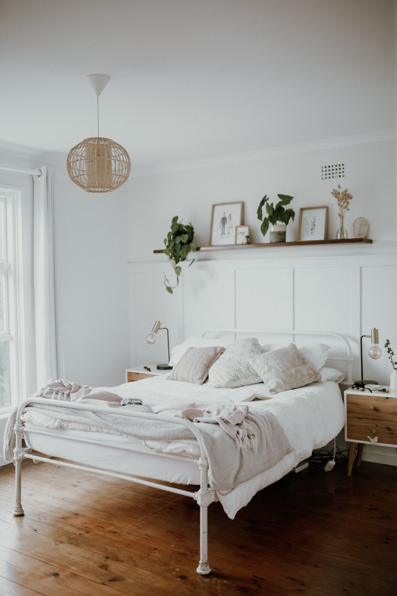 A real bedroom with white paneling, a pale painted wall, warm wood flooring, mixed bedside tables, and layered neutral bedding.